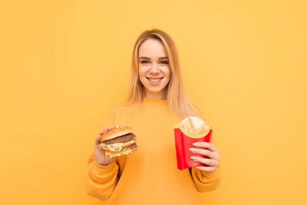 캐주얼 한 옷을 입은 행복한 학생 소녀는 햄버거와 감자 튀김을 손에 들고 미소를 지으며 노란색 배경의 카메라를 바라보고 있습니다. 색깔배경에 나쁜 음식을 가진 소녀, 고립 — 스톡 사진