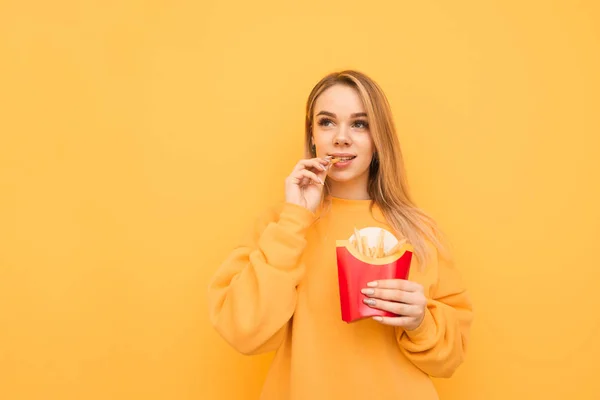 Turuncu Casual giyim sarı bir arka planda patates kızartması yeme sevimli sarışın kız portre. Güzel öğrenci patates kızartması ısırır, izole. Kopya-boşluk — Stok fotoğraf