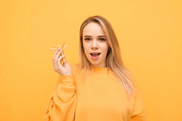 Ragazza bionda stupita con le patatine fritte mano, guardando in macchina fotografica su uno sfondo giallo, indossando una felpa arancione. Studente con una manciata di patatine fritte posa su uno sfondo giallo, volto stupito . — Foto Stock