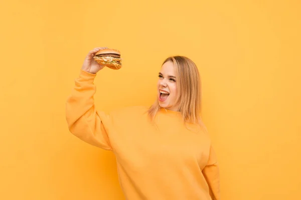노란 배경에 햄버거를 들고 패스트 푸드를 보고 기뻐하는 표현소녀의 초상화. 즐거운 소녀는 그녀의 손에 햄버거를보고 소리. 패스트푸드 컨셉. — 스톡 사진