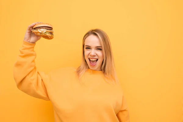 Menina alegre segura um delicioso hambúrguer na mão, olha para a câmera e se alegra, use camisola alaranjada.Menina com fome com uma má comida na mão e um rosto emocional, isolado em um fundo amarelo. — Fotografia de Stock