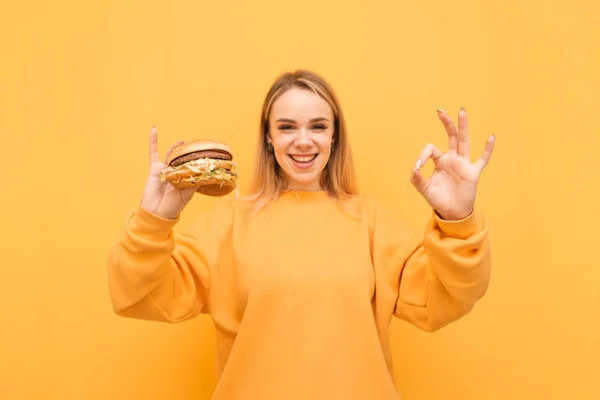 행복한 소녀는 확인 기호를 표시하고 카메라를 보고 노란색 배경에 고립 된 그의 손에 햄버거를 들고있다. 배고픈 소녀는 패스트 푸드를 먹는다. 학생은 패스트 푸드를 먹는다. 확인 기호. — 스톡 사진