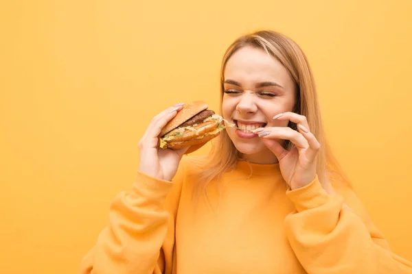 배고픈 표현 소녀는 오렌지 옷을 입고, 노란색 배경에 좋은 맛있는 햄버거를 먹는다. 소녀는 식욕을 돋우는 패스트 푸드를 먹고 즐긴다. 격리. 클로즈업 초상화 — 스톡 사진