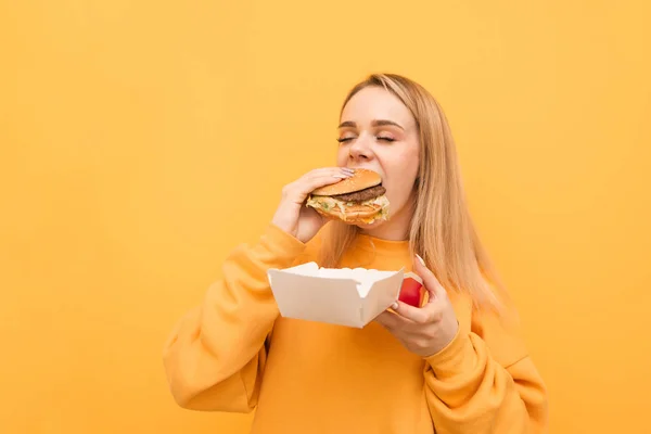 Retrato de cerca de una chica que muerde una hamburguesa con los ojos cerrados sobre un fondo amarillo, vistiendo ropa anaranjada.Chica hambrienta come un alimento dañino, sostiene el embalaje y la hamburguesa . — Foto de Stock