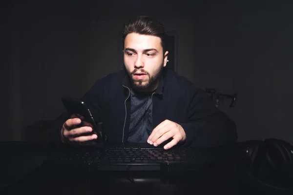 Man met een baard en in donkere kleren zit op een computer 's nachts en maakt gebruik van een smartphone. Portret van een freelancer, werkt thuis op een computer en kijkt naar het telefoon scherm. — Stockfoto