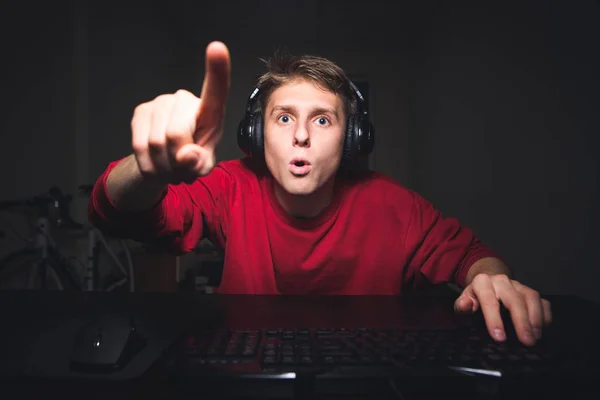 Amazed young man sitting at the table at the computer at home, with astonished face looking at the screen and showing his finger,wearing headphones.Amazed gamer gamer plays games on a computer — Stock Photo, Image
