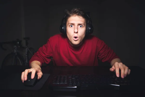 Портрет эмоционального геймера с удивленным лицом, сидящего дома на компьютере, смотрящего на экран с открытым ртом. Подросток носит красный свитер и наушники, играет в игры на компьютере дома — стоковое фото