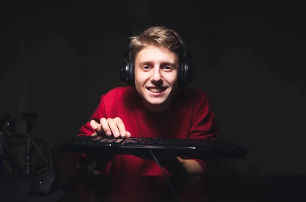 Улыбающийся геймер играет в видеоигры на фоне темной комнаты, держит в руке клавиатуру, носит наушники. Портрет счастливого подростка, использует компьютер дома, смотрит в камеру и улыбается — стоковое фото