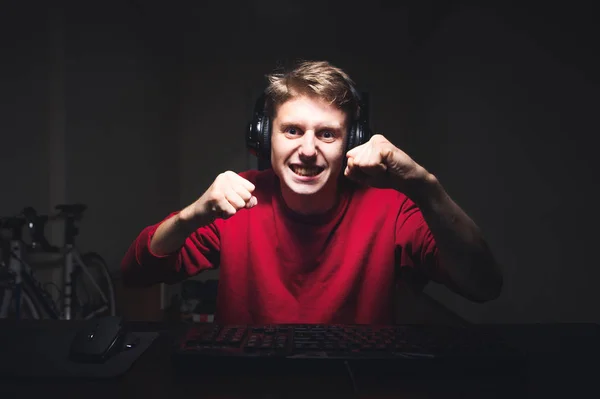 Expressieve gamer zit aan het hoofd van de computer 's nachts, kijkt naar het scherm en toont zijn vuisten. Emotionele tiener is het spelen van video games op een computer, hij is boos, zijn vuisten gebalde — Stockfoto