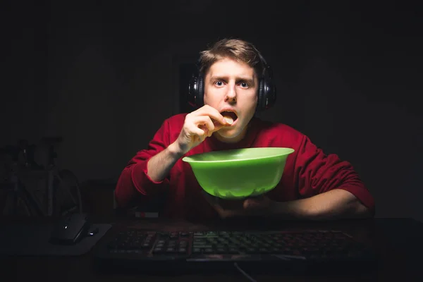 Сконцентрированный молодой человек сидит по ночам в комнате, смотрит фильмы на компьютере и ест зеленую закуску блюдо. Парень ест чипсы и выглядит сосредоточенным на экране компьютера на фоне темного Ру — стоковое фото