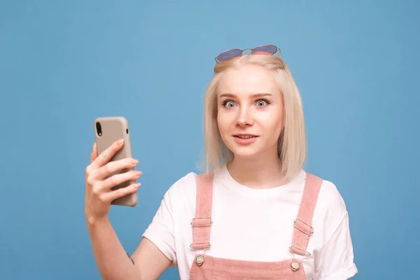 Menina emocional com um smartphone em suas mãos em um fundo azul olha para a câmera com espanto. Retrato de close-up de uma adolescente surpresa com um smartphone em sua mão. Isolados . — Fotografia de Stock