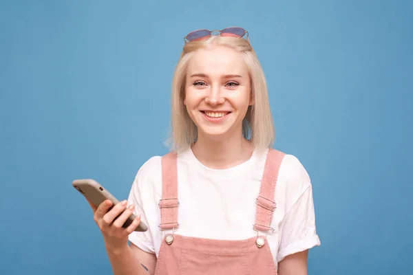 Retrato de close-up de uma menina feliz em um fundo azul, segura um smartphone em suas mãos, olha para a câmera e sorri. Menina adolescente positivo com smartphone na mão é isolado no fundo azul — Fotografia de Stock