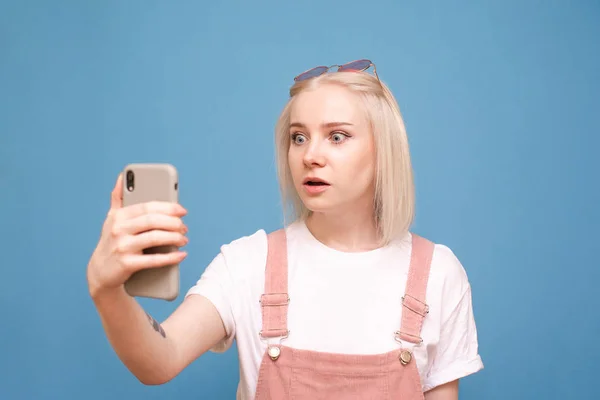 Retrato de uma menina bonito surpreso com um smartphone na mão, com o rosto espantado olhando para a tela de um telefone em um fundo azul.Menina expressiva usa um smartphone, emoção surpresa. — Fotografia de Stock