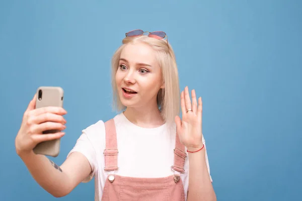 Menina doce com um telefone falando em uma chamada de vídeo no smartphone, olhando para a tela do telefone com rosto surpreso em um fundo azul. Adolescente menina faz selfie em um fundo azul . — Fotografia de Stock