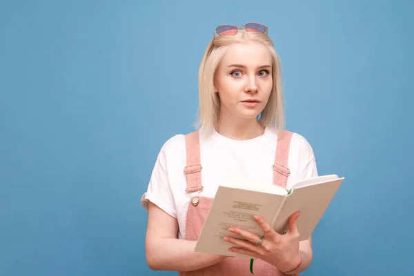 Συναισθηματικό κορίτσι με ένα βιβλίο με χέρια που στέκεται σε μπλε φόντο, κοιτάζοντας μέσα στην κάμερα με έκπληκτος πρόσωπο. Χαριτωμένο έφηβος κορίτσι κρατά ένα βιβλίο στα χέρια της, φοράει γυαλιά, ένα λευκό μπλουζάκι και ένα φόρεμα — Φωτογραφία Αρχείου