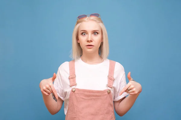 Menina loira surpreso em roupas bonitos fica em um fundo azul, mostrando os dedos para baixo e olhando para camera.Teenage menina com um rosto surpreso, mostrando as mãos para baixo, isolado em um fundo azul — Fotografia de Stock