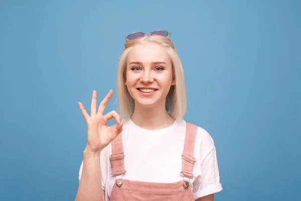 Happy teen girl mostra un segno OK guarda la fotocamera e sorride, indossa un abbigliamento casual carino e occhiali da sole, isolato su uno sfondo blu. Ritratto di un adolescente felice . — Foto Stock