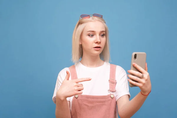Retrato de una chica linda sorprendida muestra el dedo en el teléfono inteligente en su mano y mira la pantalla.Chica adolescente emocional en ropa linda utiliza un teléfono inteligente con una cara sorprendida en un fondo azul — Foto de Stock