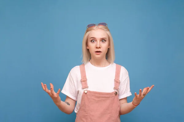 Shocked girl teenager is isolated on a blue background and looks surprised face in the camera, wears a white T-shirt and a pink sundress.Emotionally blonde girl with a shocking face, studio portrait