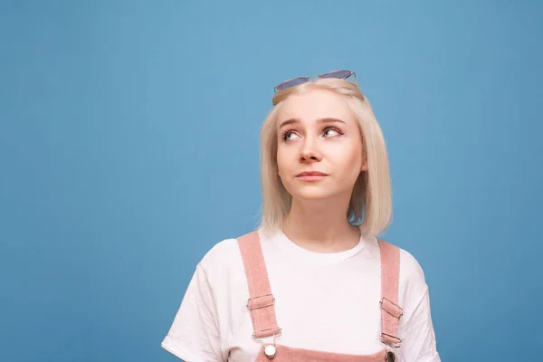 Menina bonito em um vestido casual brilhante, vestindo uma camiseta branca, óculos de sol rosa e vestido, olha para o lugar vazio com um rosto engraçado. Menina adolescente positivo em um fundo azul olha para o lado — Fotografia de Stock