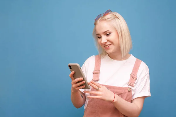 Menina sorridente adorado com cabelo claro, usa um telefone em um fundo azul. Senhora feliz em roupas brilhantes com um smartphone em sua mão é isolado em um fundo azul . — Fotografia de Stock