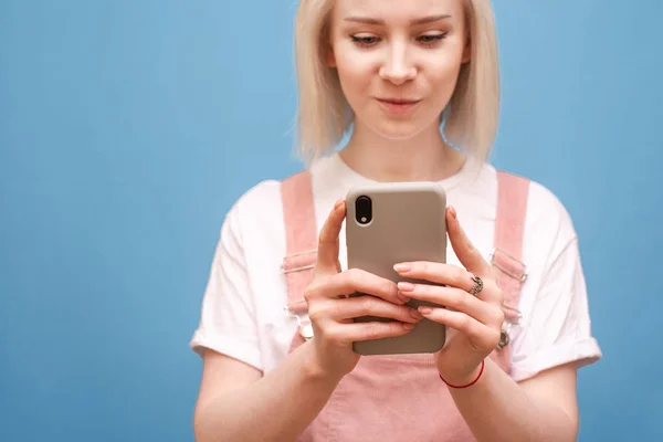 Smartphone en manos de una adolescente. Foto de cerca de una chica rubia en ropa linda utiliza un teléfono inteligente sobre un fondo azul, el enfoque en el teléfono en las manos de la chica. Espacio de copia — Foto de Stock