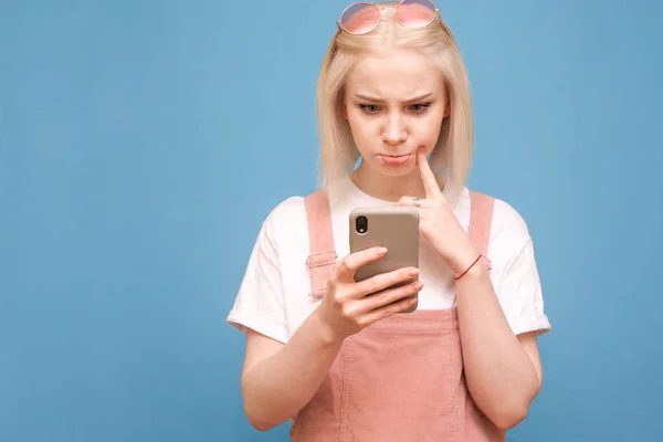Chica rubia divertida en ropa linda con una cara reflexiva utiliza un teléfono inteligente sobre un fondo azul, el enfoque en el teléfono en las manos de la chica. Espacio de copia — Foto de Stock
