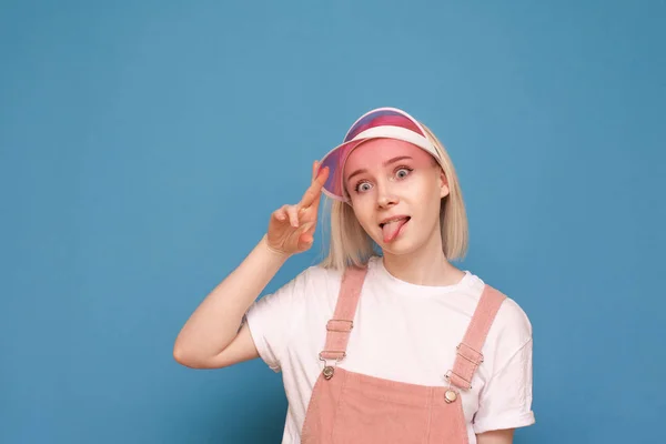 Retrato de una chica divertida en un vestido bastante casual, con una gorra rosa y haciendo una cara divertida sobre un fondo azul. Divertido adolescente chica poses en la cámara, aislado primer plano retrato . —  Fotos de Stock