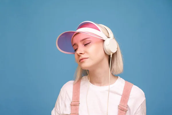 Retrato de uma loira atraente com um boné rosa e uma camiseta branca fica no fundo de uma parede azul e ouve música nos fones de ouvido com olhos fechados, obtém prazer da música . — Fotografia de Stock