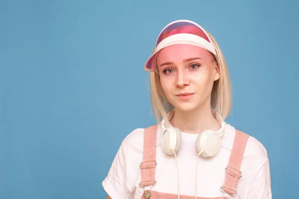 Bella ragazza con le cuffie e un berretto rosa è su uno sfondo blu e guarda la fotocamera, un ritratto da vicino. Ritratto di un'adolescente che posa sullo sfondo di un muro blu. Isolato . — Foto Stock