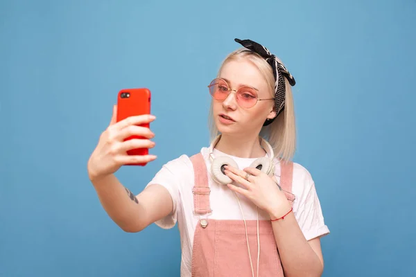 Stijlvol meisje in een koptelefoon en heldere kleren maakt een selfie op een smartphone op een blauwe achtergrond, kijkend naar het scherm. Tiener neemt een selfie op een blauwe achtergrond. — Stockfoto