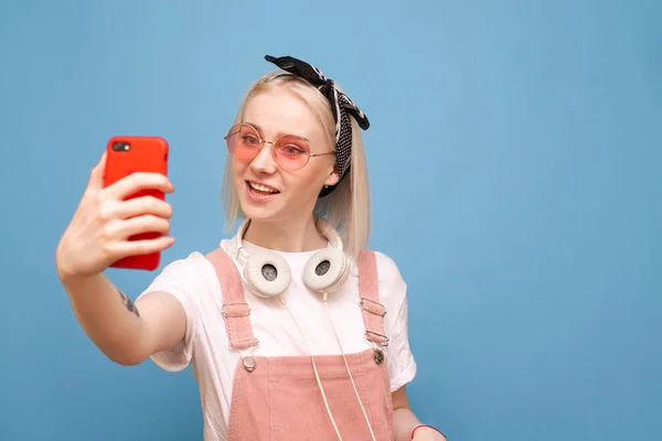 Menina feliz em roupas brilhantes e fones de ouvido elegantes em pé sobre um fundo azul faz selfie e sorrisos. Menina positiva nos óculos de sol e um smartphone na mão é isolado em um fundo azul — Fotografia de Stock