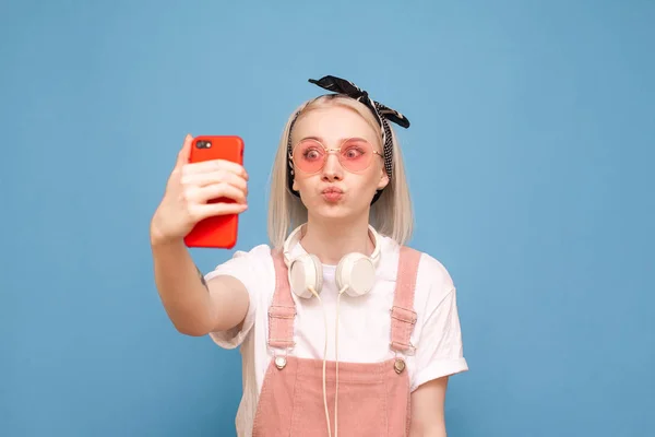 Chica divertida en ropa linda toma una selfie con una cara divertida sobre un fondo azul. Divertida adolescente chica hace selfie en un teléfono inteligente, con ropa brillante y gafas de sol de color rosa — Foto de Stock