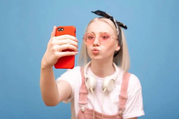 Ritratto ravvicinato di una ragazza divertente in occhiali rosa brillante prende un selfie su uno smartphone con una faccia divertente su uno sfondo blu. Focus sullo smartphone . — Foto Stock