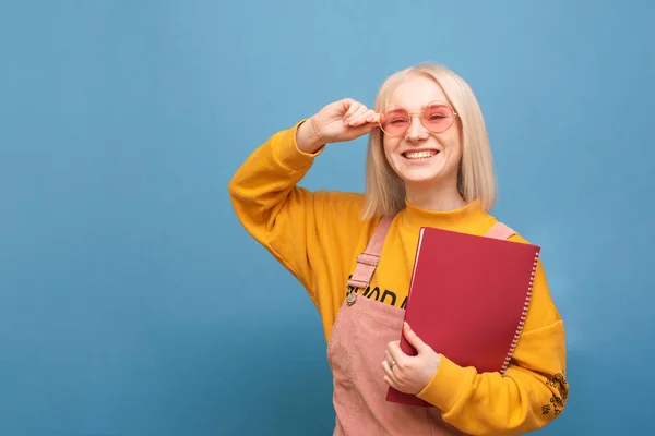 Portret van een gelukkige student meisje in roze bril en een Kladblok in haar hand staat op een blauwe achtergrond, kijkt naar de camera en glimlacht. Stijlvol meisje met boeken in haar handen glimlachend, geïsoleerd. — Stockfoto