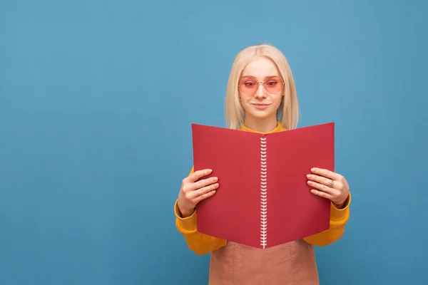 Πορτρέτο ενός αστεία ξανθιά σε ροζ γυαλιά σε ένα μπλε φόντο, κρατά ένα κόκκινο σημειωματάριο στα χέρια του και διαβάζει. Χαρούμενο κορίτσι μαθητής μαθαίνει. Απομονωμένες. Χώρος αντιγραφής — Φωτογραφία Αρχείου