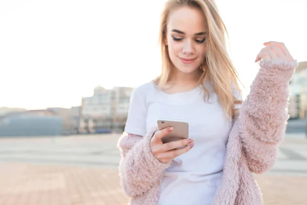 Девушка в розовом пальто пользуется смартфоном на улицах. — стоковое фото