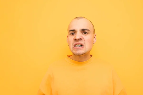 Retrato de um jovem emocional com grillz em seus dentes, vestindo uma roupa casual brilhante e faz um rosto malicioso em um fundo amarelo. Malvado cara elegante com uma cabeça careca e uma cerda — Fotografia de Stock