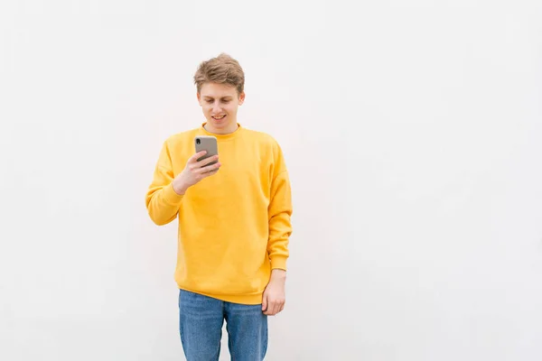Smiling teenager in a stylish casual clothing is standing on the background of a white wall, using a smartphone and smiling. Street portrait of a stylish boy with a smarthon in his hands. — Stock Photo, Image