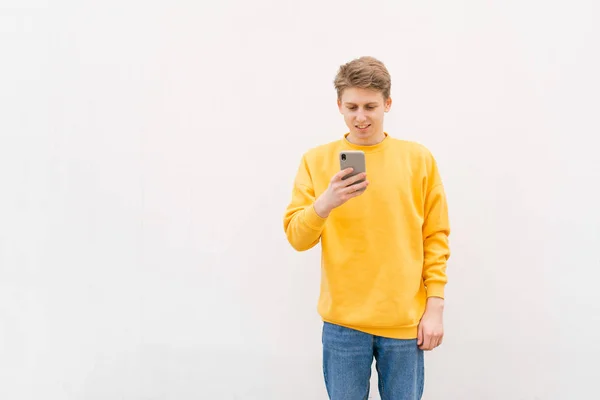 Retrato de un estudiante usando un teléfono inteligente en el fondo de una pared blanca, mirando la pantalla del teléfono inteligente y sonriendo. Retrato de un joven con ropa brillante sobre un fondo blanco. Aislado . —  Fotos de Stock