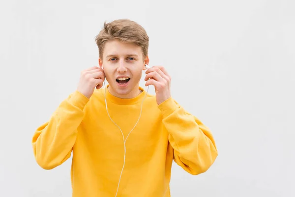 Retrato de un joven emocional con una sudadera amarilla en el fondo de una pared blanca en los auriculares, se ve sorprendido en la cámara, boca abierta.Adolescente con estilo escucha música sobre un fondo blanco —  Fotos de Stock