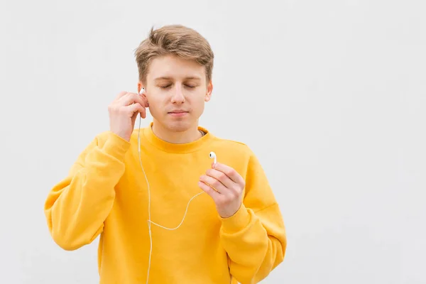 Retrato de primer plano de un joven parado en el fondo de una pared blanca, inserta un auricular con cable en sus oídos con los ojos cerrados y disfruta de la música.Tipo con auriculares en el fondo de una pared blanca — Foto de Stock