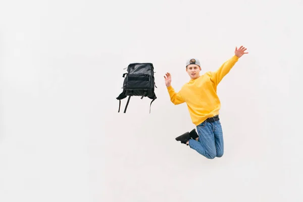 Elegante giovane uomo in un salto con uno zaino su uno sfondo bianco con indosso un abbigliamento casual luminoso. Studente con uno zaino in volo sullo sfondo di un muro bianco. Isolato. Copyspace . — Foto Stock