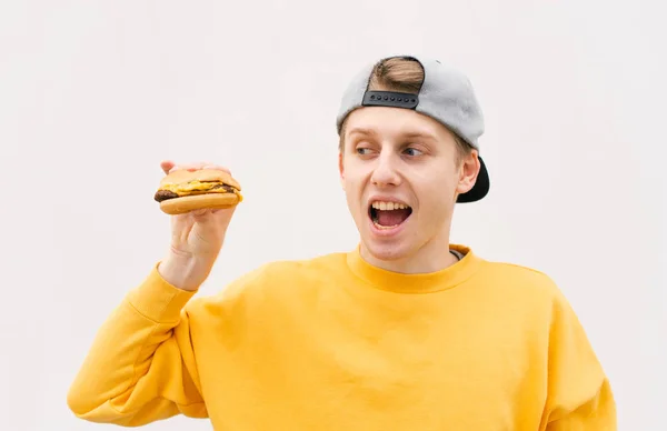 Foto de cerca de un tipo positivo en un vestido casual con un sándwich en la mano contra un fondo blanco de la pared, mirando al lado de la comida rápida y sonriendo. Copyspace. Estudiante y comida rápida — Foto de Stock
