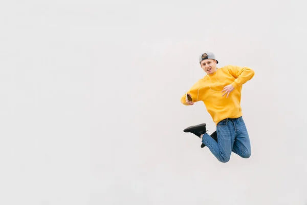 Estudiante positivo saltando en el fondo de una pared blanca, usando una ropa casual con estilo, escucha música en los auriculares, sostiene un teléfono inteligente en la mano. Chico feliz saltando sobre fondo claro —  Fotos de Stock