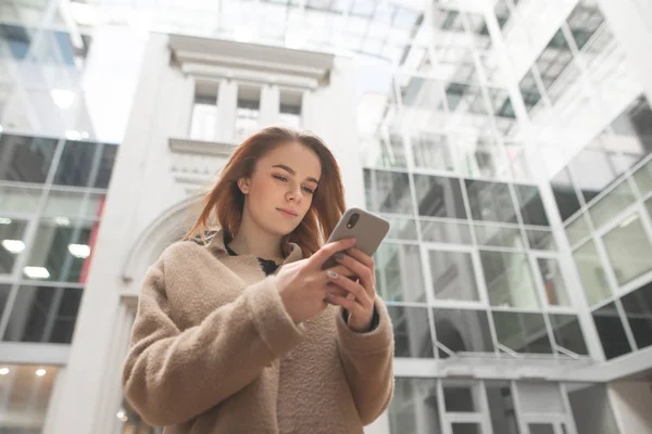 Bir ceket giyen ciddi genç iş kadını, ekran odaklı panoramik pencereler ile modern mimarinin arka planda bir akıllı telefon kullanır. Elinde bir akıllı telefon ile bir kızın kentsel portresi — Stok fotoğraf