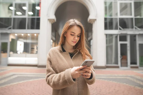 Attractive stylish woman in a coat,stands on the background of modern architecture and uses a smartphone.Urban portrait of a beautiful woman with a smartphone in hand,looking at the screen and smiling — Stock Photo, Image