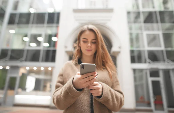 Stylish girl uses a smartphone on the street, focuses on the phone screen, against the background of modern architecture, the focus on the smartphone. Girl writes a message on phone. — Stock Photo, Image