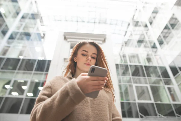 Bej bir ceket güzel kız, elinde bir akıllı telefon tutan, binanın kentsel arka plan önünde duruyor, telefon ekranına bakar. Şık bayan bir akıllı telefon kullanır — Stok fotoğraf