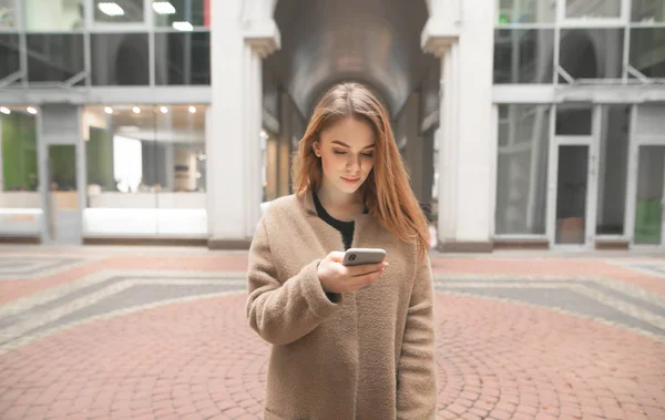 Attractive lady blonde wears a coat, uses a smartphone on the urban background. Stylish girl writes a message on a smartphone, uses the internet on the background of a modern building. — Stock Photo, Image